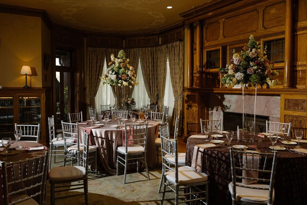 Elegant dining room with round tables set for a banquet, decorated with tall flower centerpieces, surrounded by silver chairs, and featuring a wood-paneled wall with a marble fireplace.