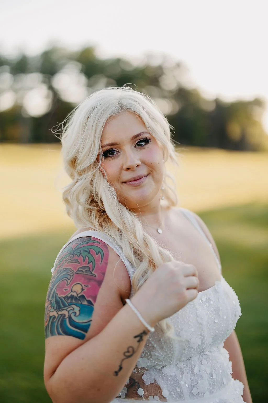 A woman with blonde hair styled in loose waves, wearing a white dress with floral appliqué, standing outdoors in a field with trees in the background, smiling softly at the camera.