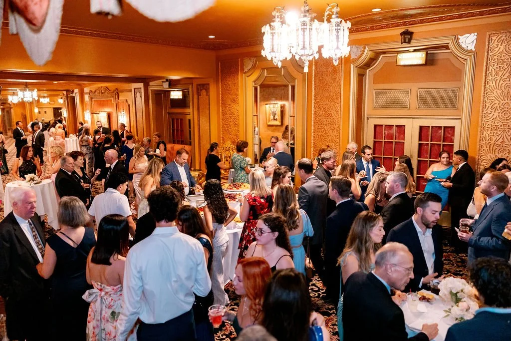 Crowd of people at a formal event or reception in an elegant, ornately decorated ballroom with warm lighting and chandeliers.