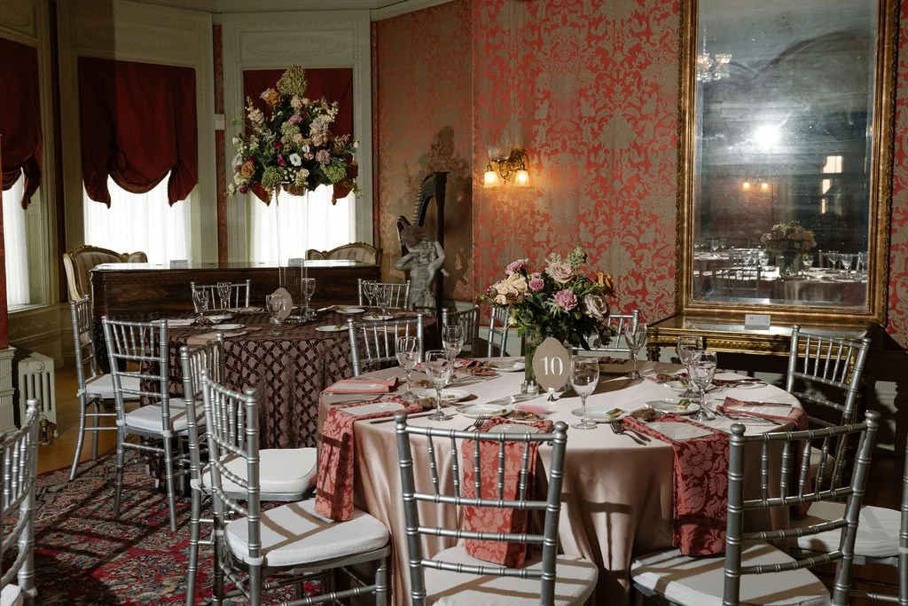 Elegant dining room with round tables set for a formal event, floral centerpieces, silver chairs, and red patterned wallpaper.