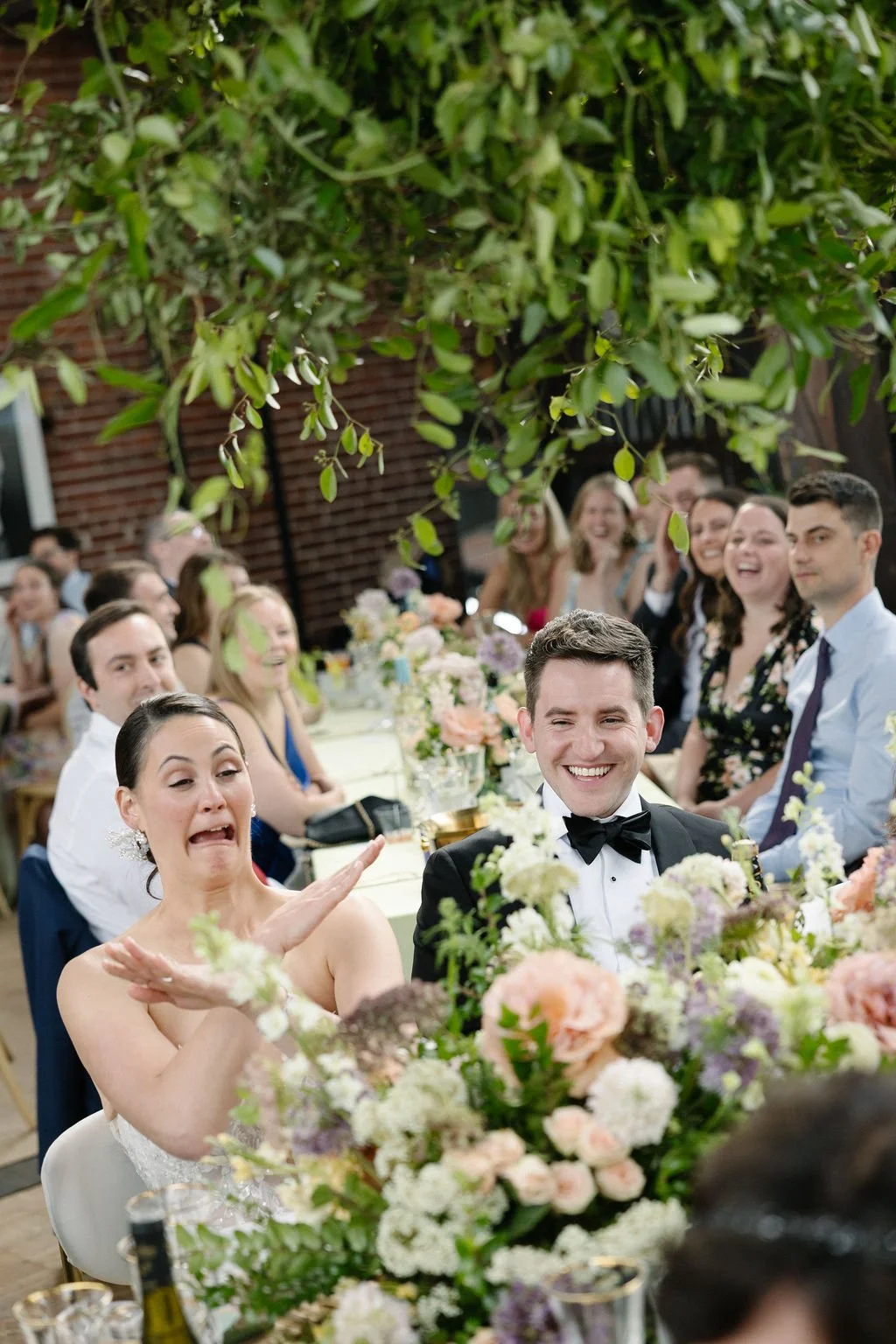 Guests at a wedding reception sitting at a long table decorated with flowers, with a large leafy plant hanging above them, smiling and laughing