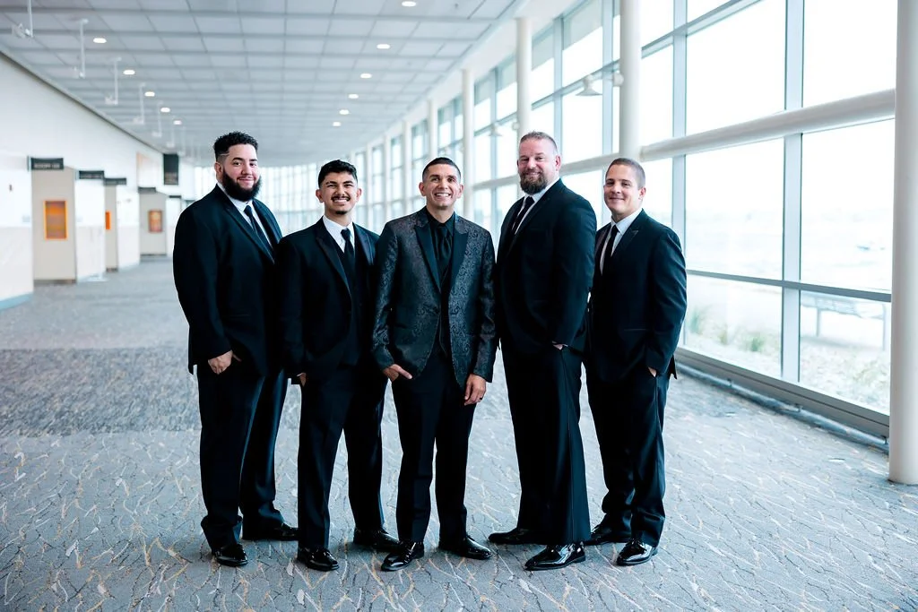 Group of five men in suits standing in an airport terminal with large windows and natural light.
