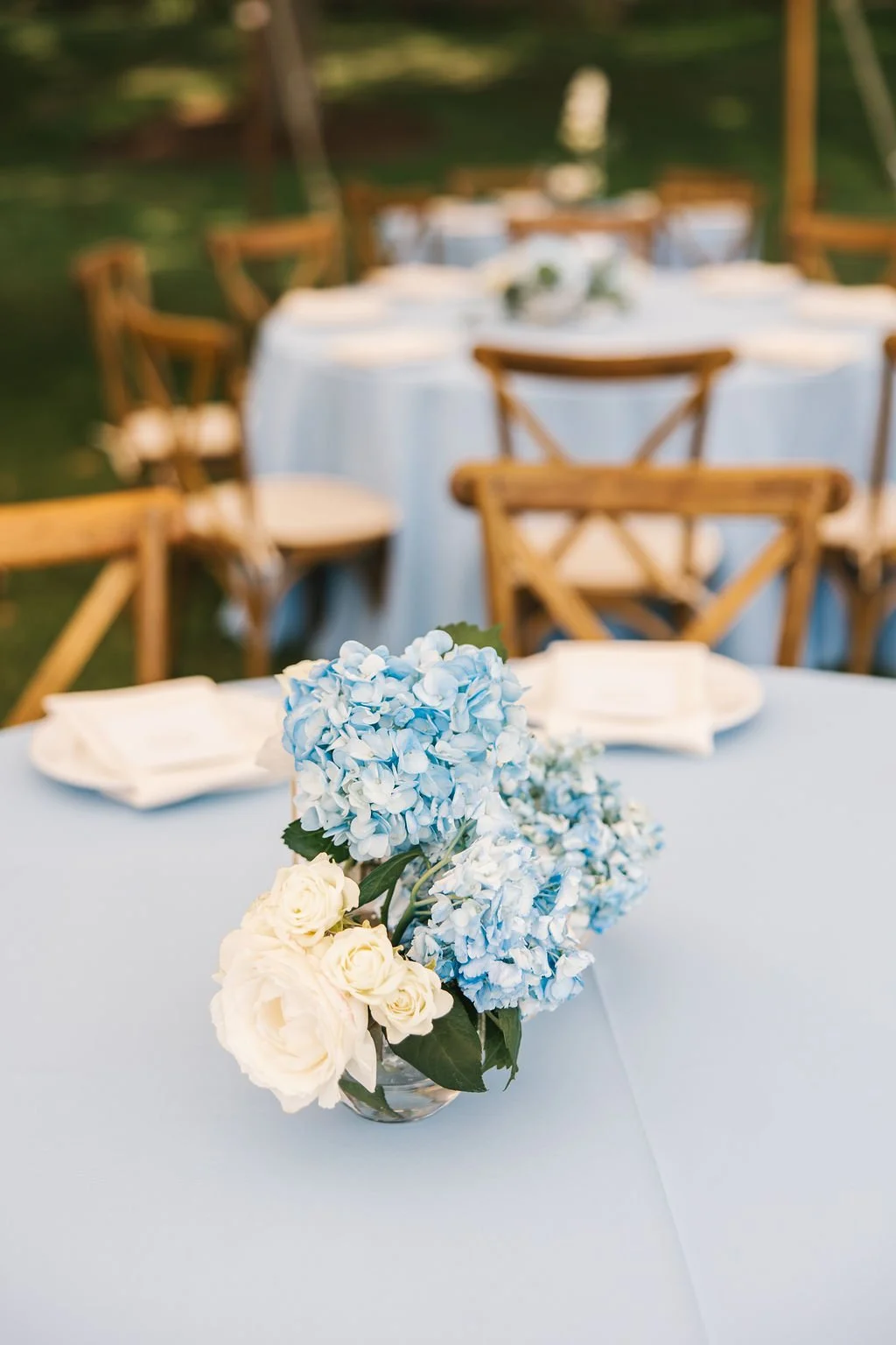 A table decorated with a bouquet of white and blue hydrangeas and roses, set with white napkins, in an outdoor setting with wooden chairs and other tables in the background.