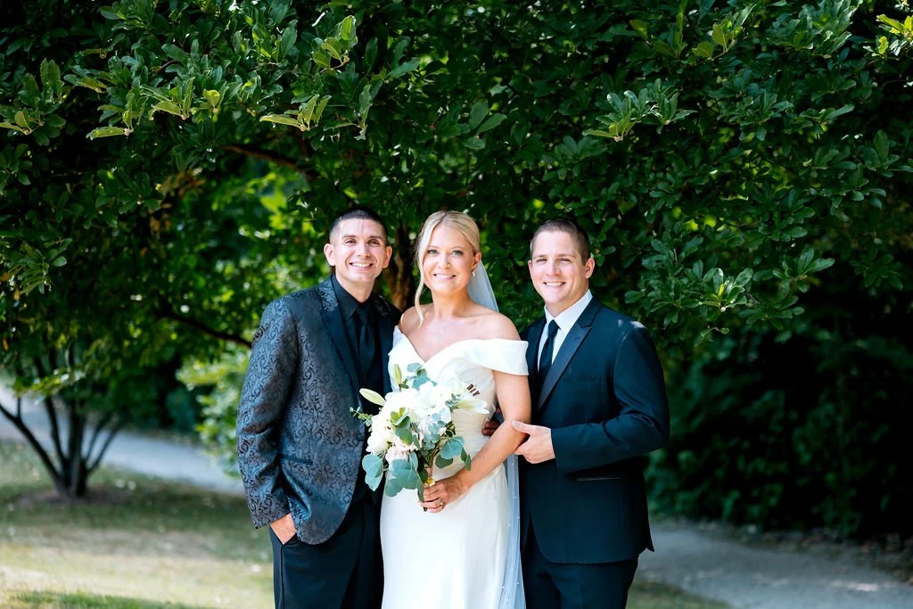 A bride in a white wedding gown holding a bouquet, standing with two men in suits outdoors against a green leafy background.