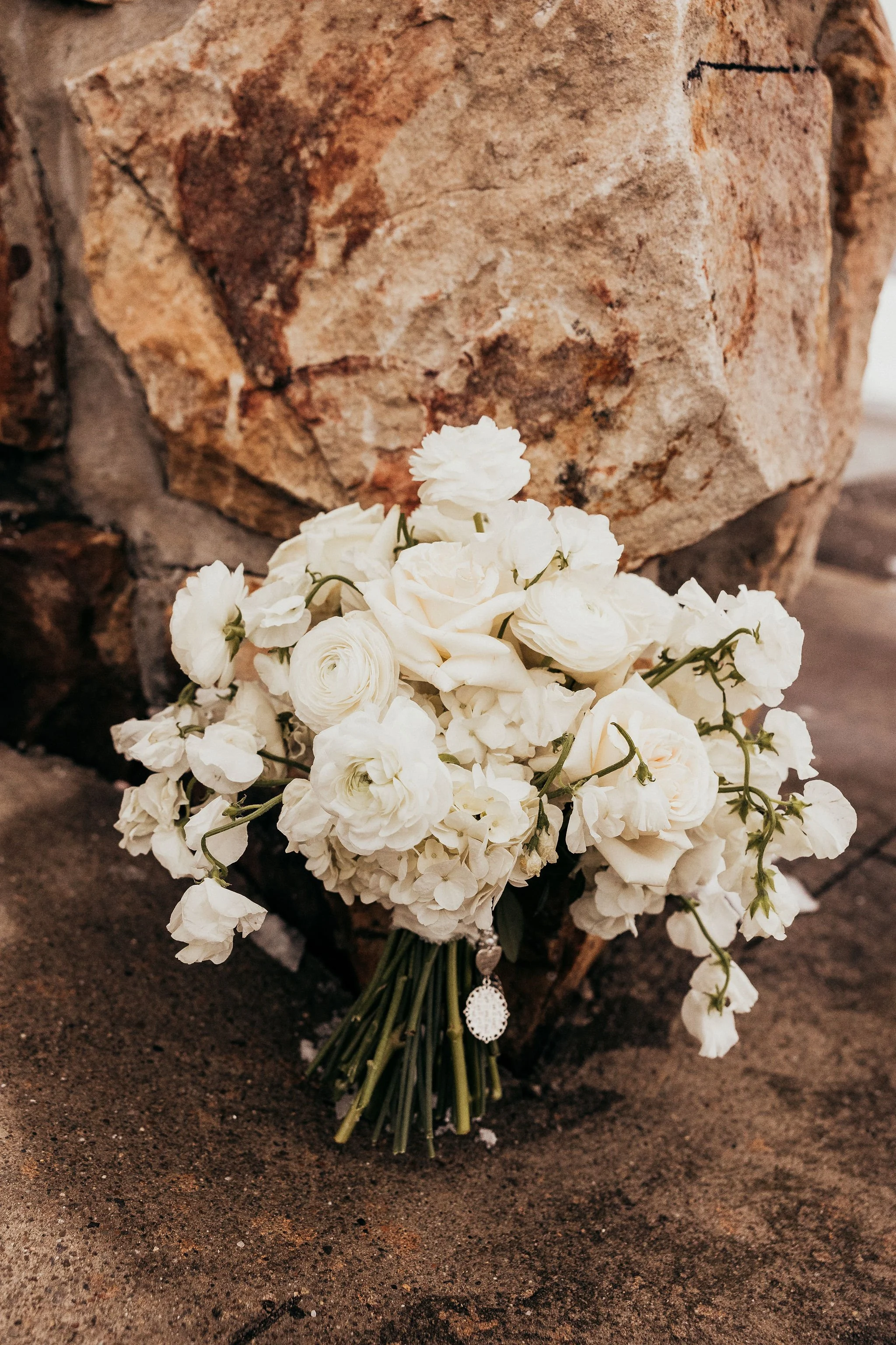 A bouquet of white flowers resting on a brown surface in front of a large, rough, beige and reddish-brown rock.