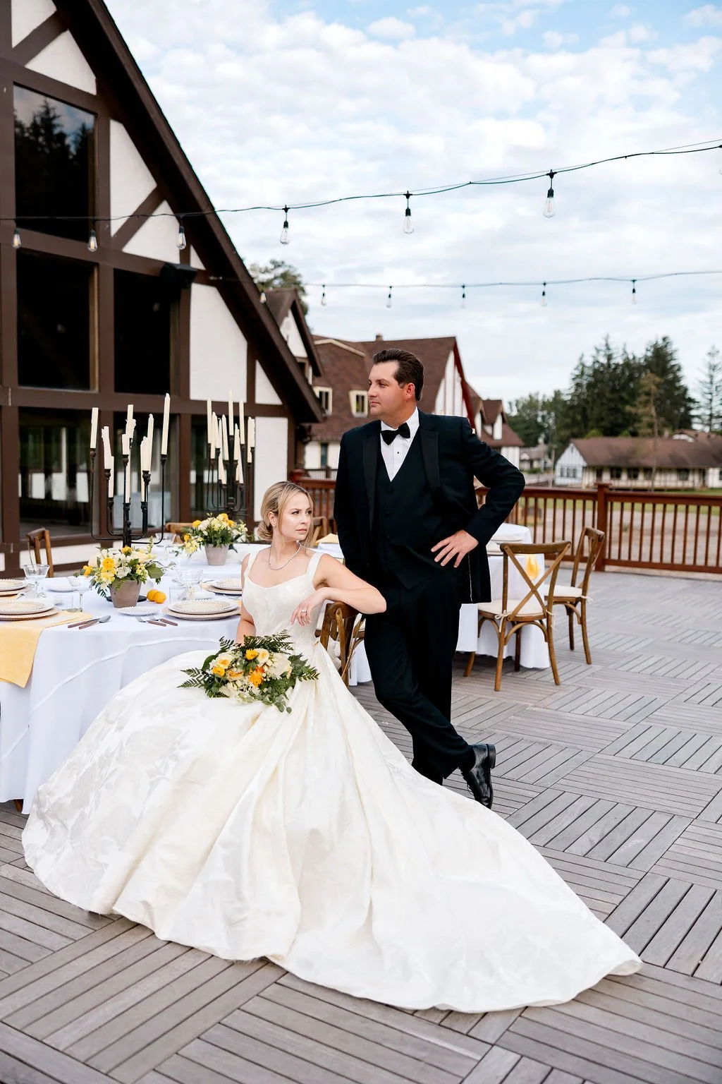 A bride in a white wedding gown with floral bouquet sitting at a table, a groom in a tuxedo standing nearby on a wooden deck outdoors, under string lights, with decorated tables and houses in the background.