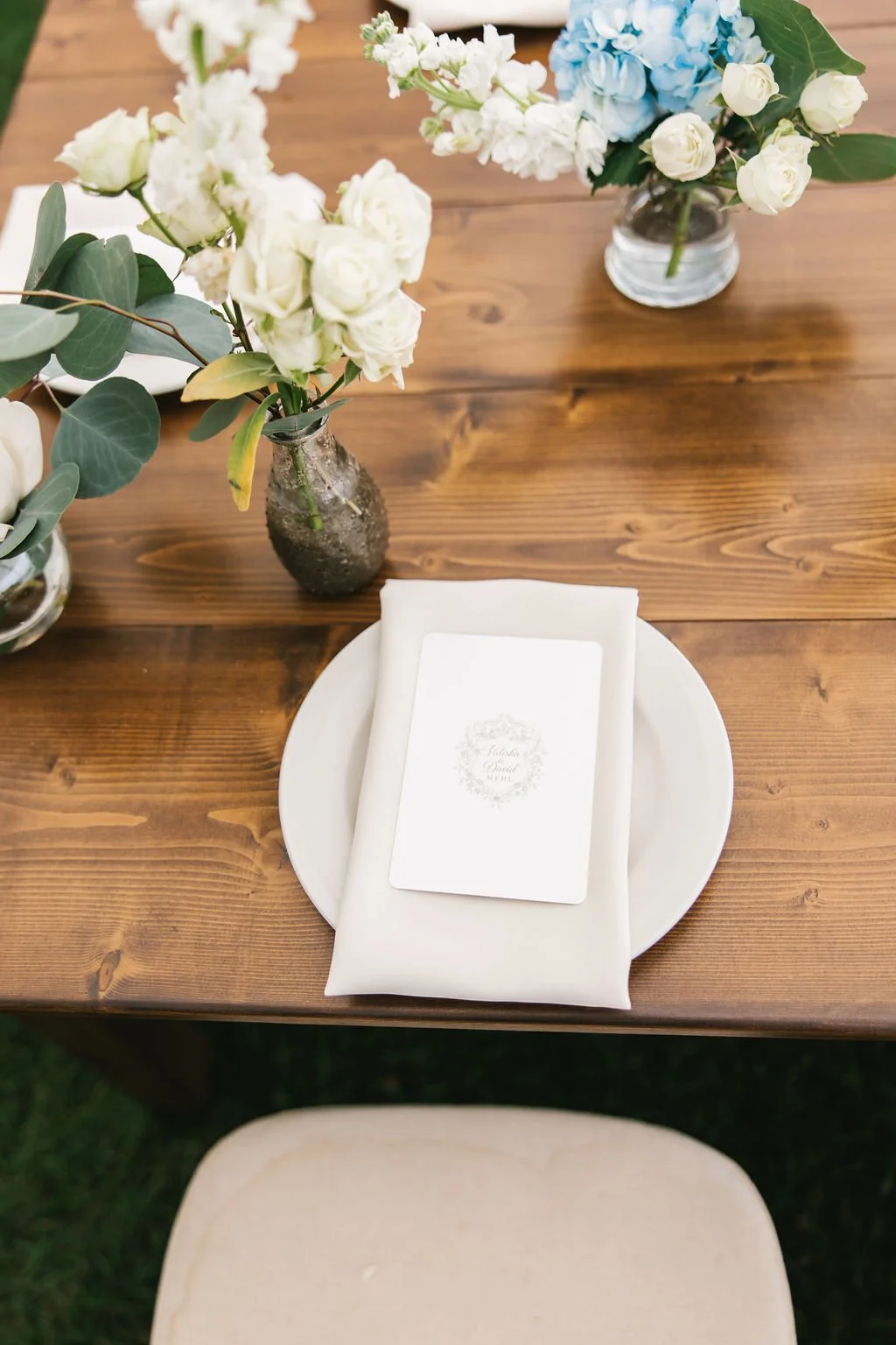 A wooden table with three small vases of white and blue flowers, a white plate with a folded white napkin, and a guest card on top of the napkin.