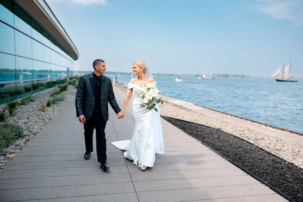 A newlywed couple walking hand in hand on a waterfront promenade, with the bride holding a bouquet of white flowers, and boats sailing on the water in the background.