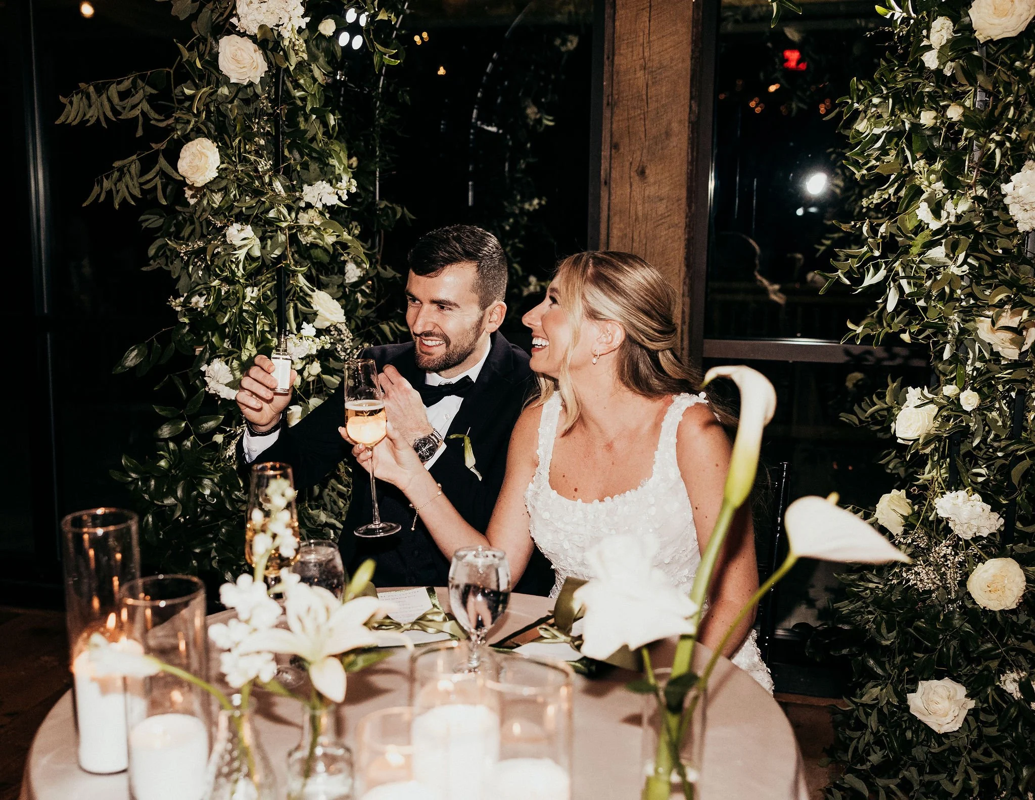 A newlywed couple, a man and a woman, sitting at a wedding reception table decorated with white flowers, holding glasses of champagne, and smiling at each other. The setting is elegant with floral arrangements and warm lighting.