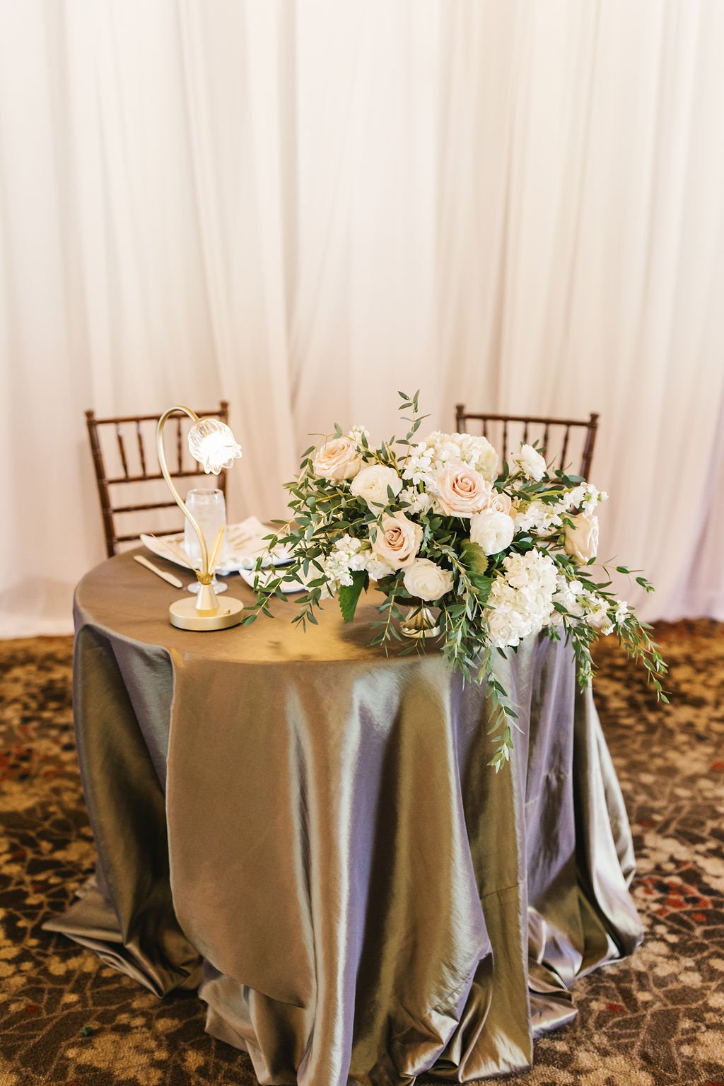 A round table with a gold-colored satin tablecloth, a floral centerpiece of white roses and hydrangeas, set with glassware and napkins, with two chairs in the background and a small lamp on the table.
