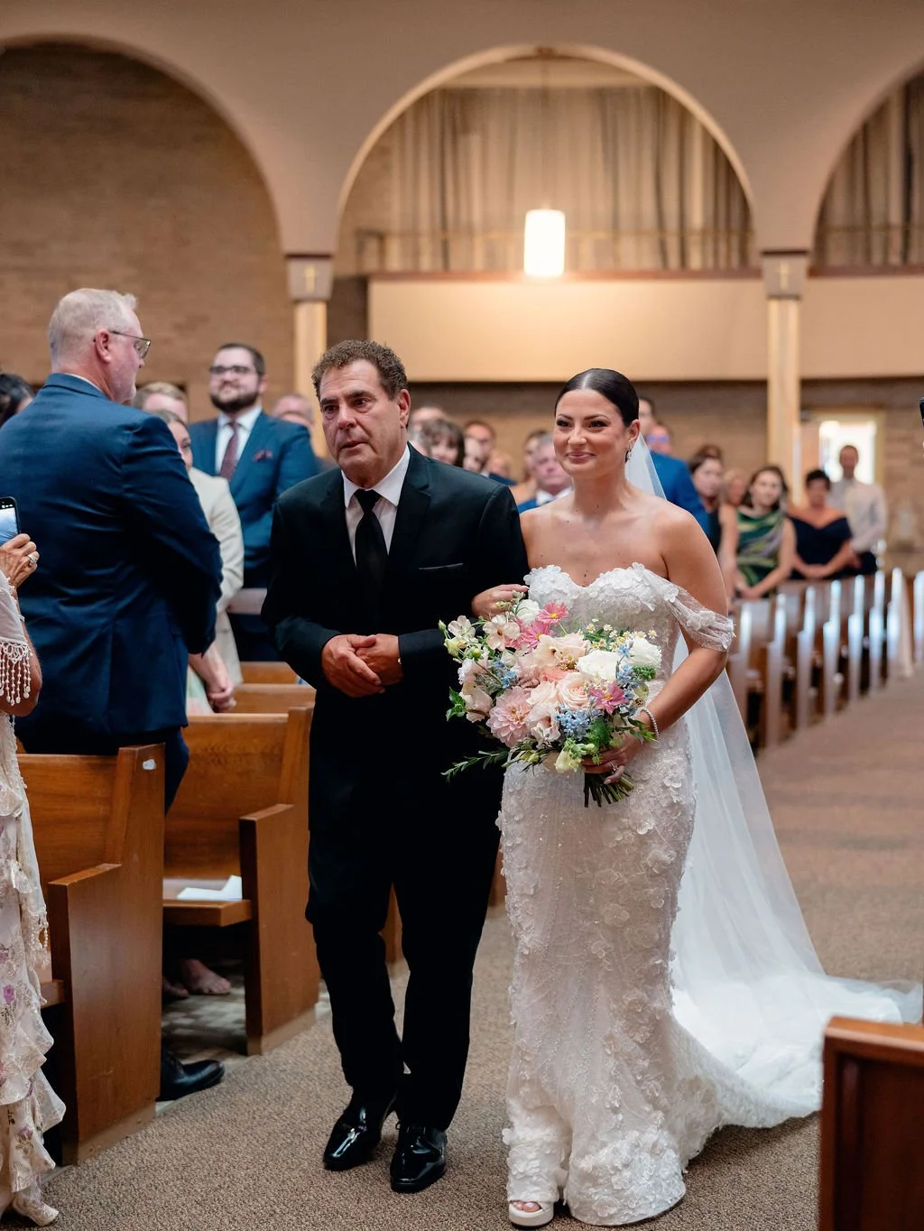 A bride walking down the aisle with her father, holding a bouquet of flowers, during her wedding ceremony in a church filled with guests.