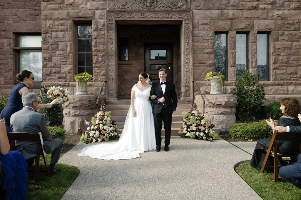 Couple in wedding attire walking down the aisle after their wedding ceremony outside a stone building with floral arrangements.