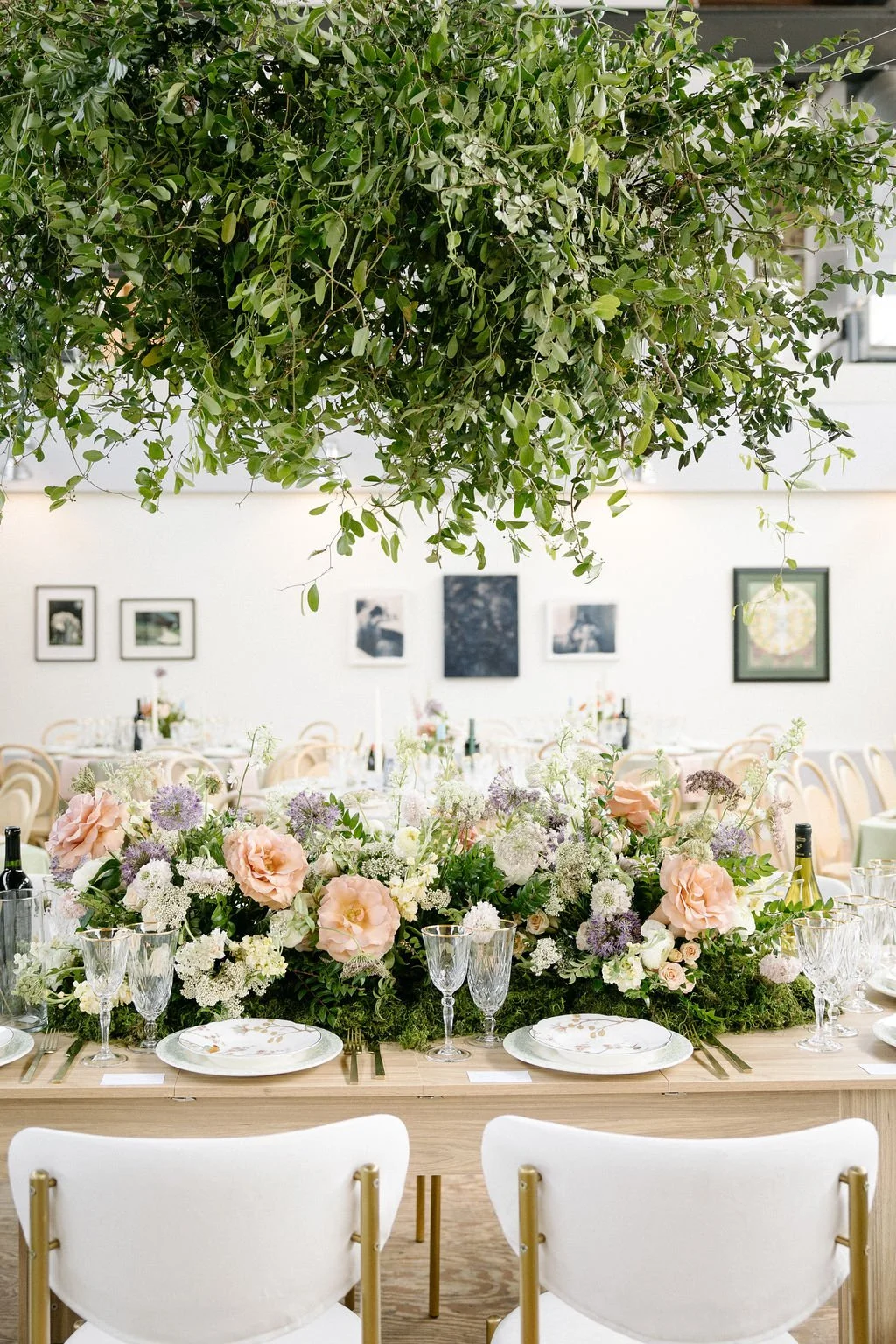 Elegant indoor event space with a long table decorated with a large floral centerpiece of pink and white flowers, surrounded by glassware and table settings, under a lush green hanging plant ceiling, with framed artwork on the white wall in the backg