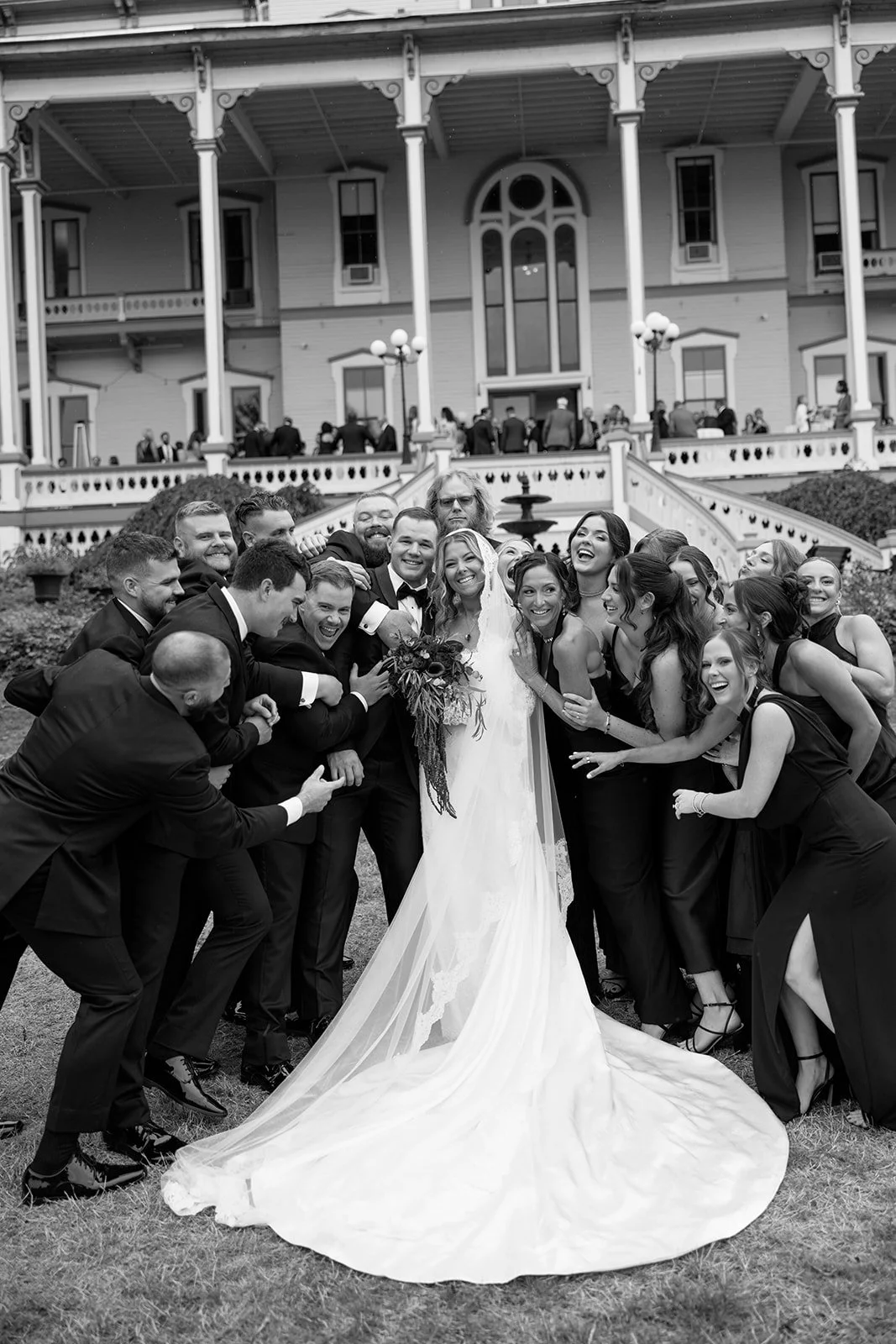 A bride and groom with a large group of people, all dressed in formal attire, posing for a photo outside a large, ornate house.