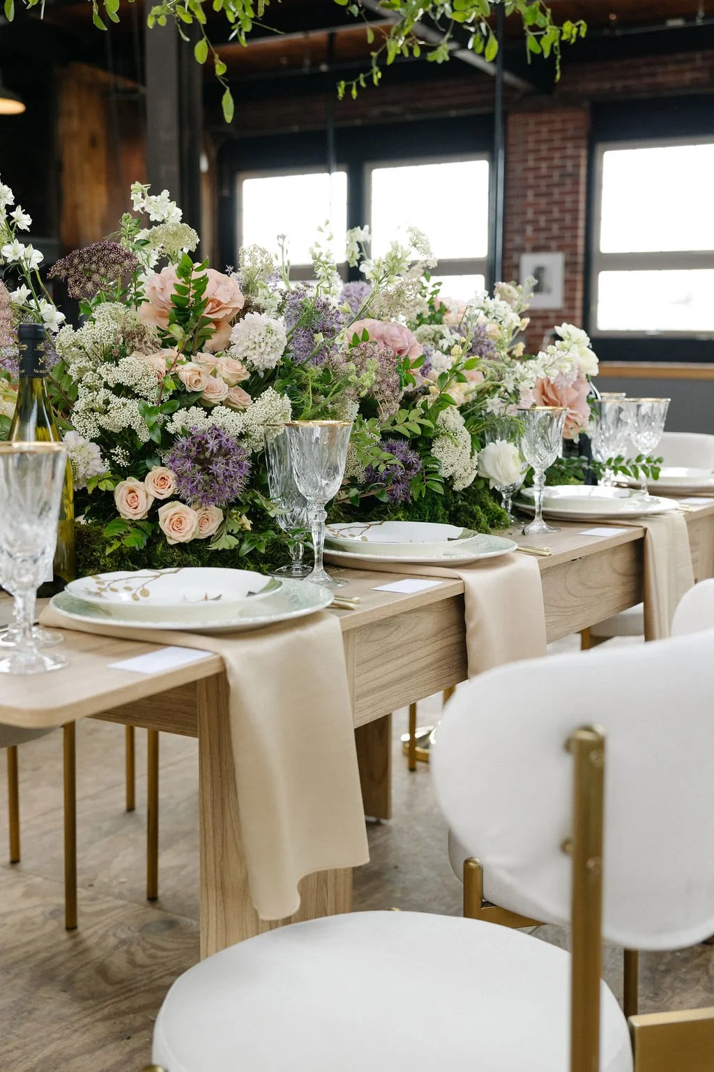 A beautifully decorated dining table with a large floral centerpiece, wine glasses, porcelain plates, and beige table runners set in a rustic venue with brick walls and large windows.