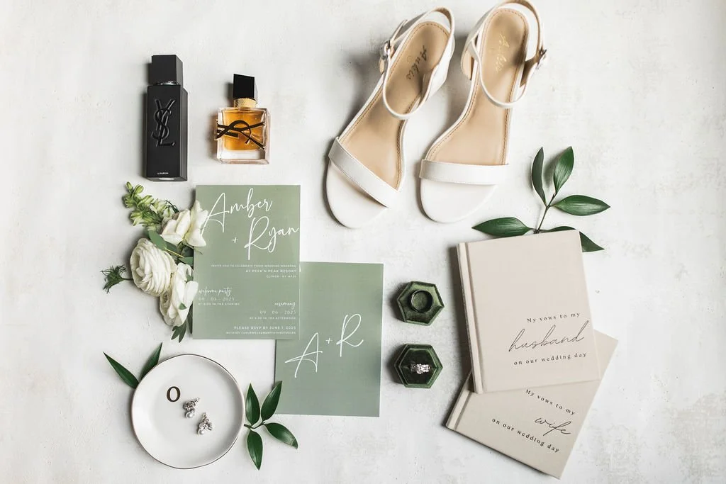 Wedding invitation suite with rings, shoes, perfume, and floral accessories arranged on a white surface.