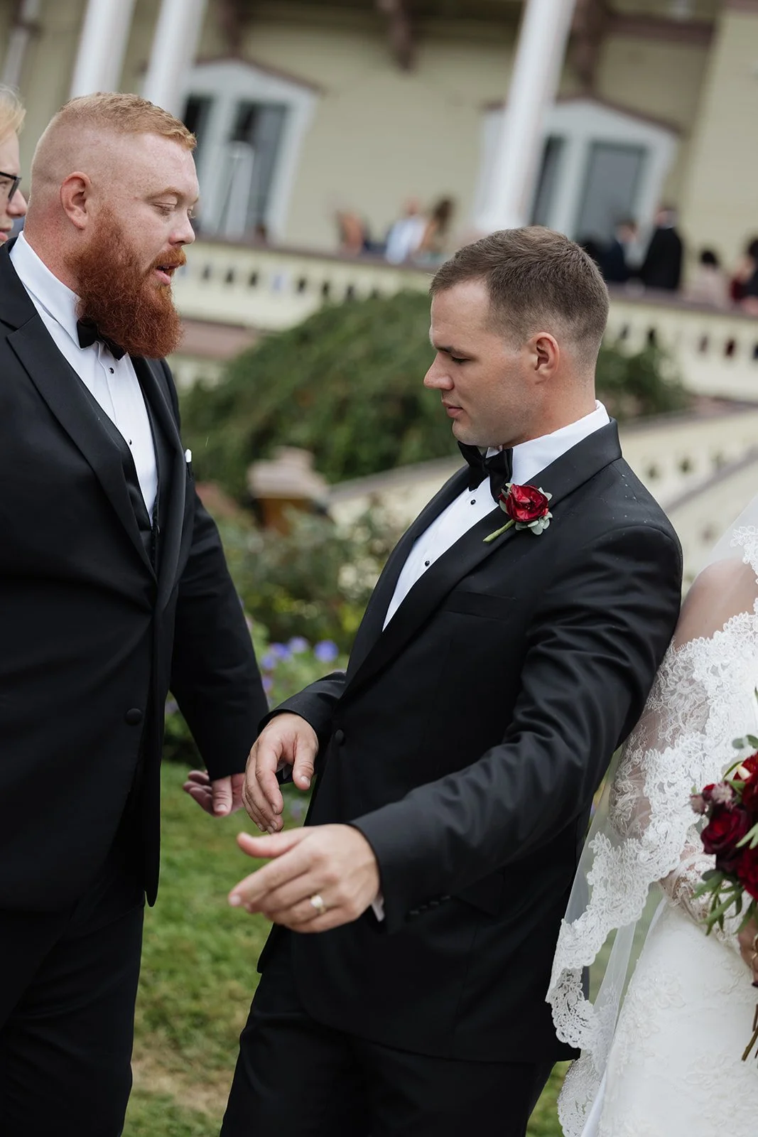 A groom with short hair and a shaved side, wearing a black tuxedo with a white shirt and a black bow tie, standing outside during a wedding ceremony, holding hands with another man with a red beard, who is also dressed in a black tuxedo with a white 