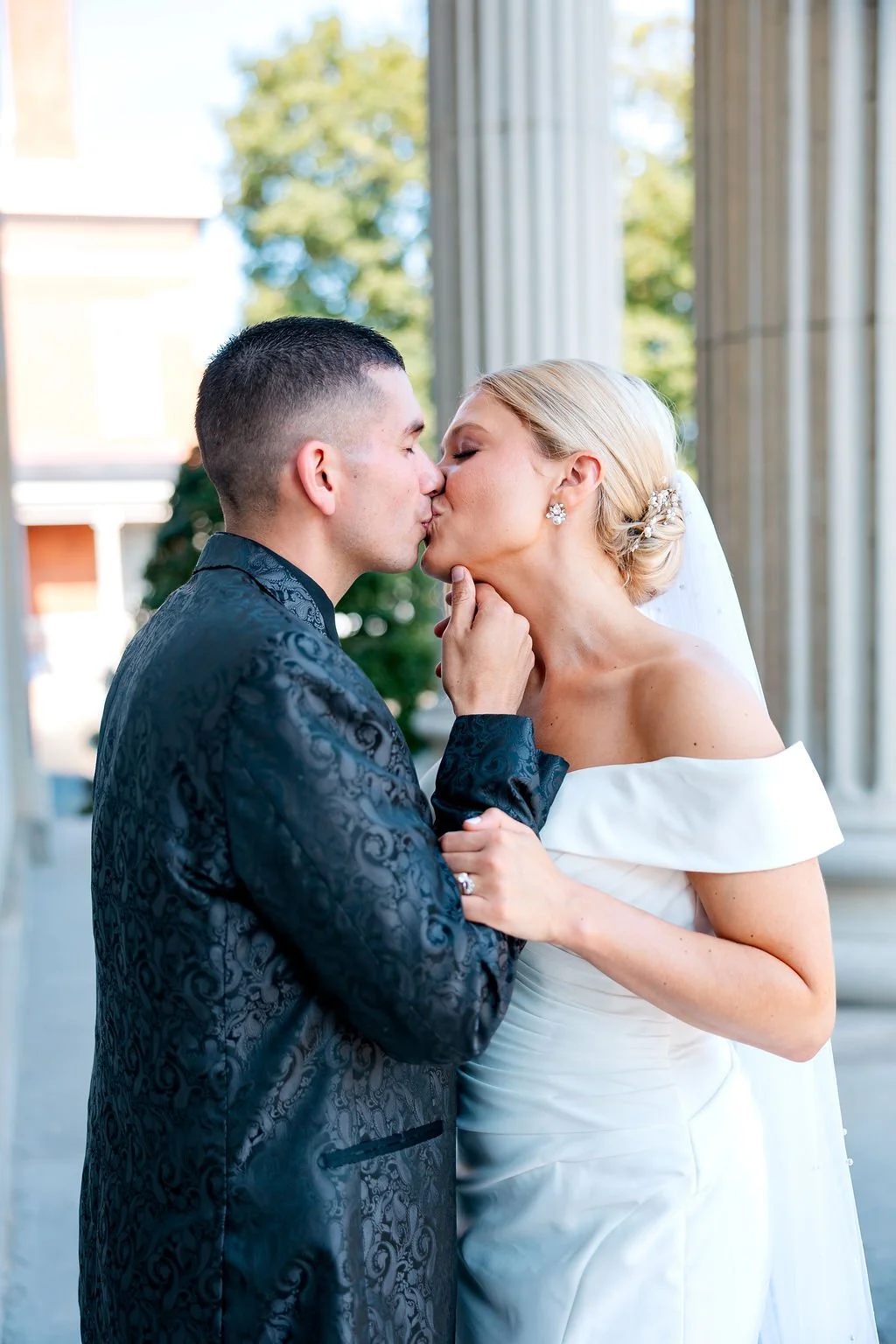 A bride and groom sharing a kiss outdoors, with the bride wearing a white off-the-shoulder wedding dress and the groom in a dark patterned suit.