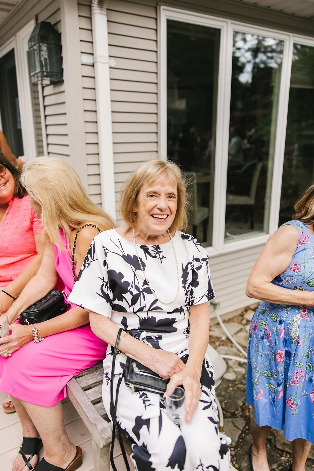 smiling elderly woman in a black and white floral dress sitting on a wooden bench at an outdoor gathering with other women, one in a pink dress and another in a blue floral dress, near a house with gray siding and large windows