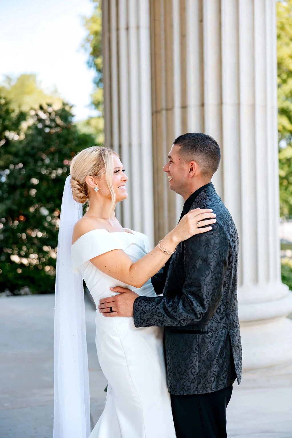 A bride and groom smiling at each other during their wedding outside, with large classical columns and greenery in the background.
