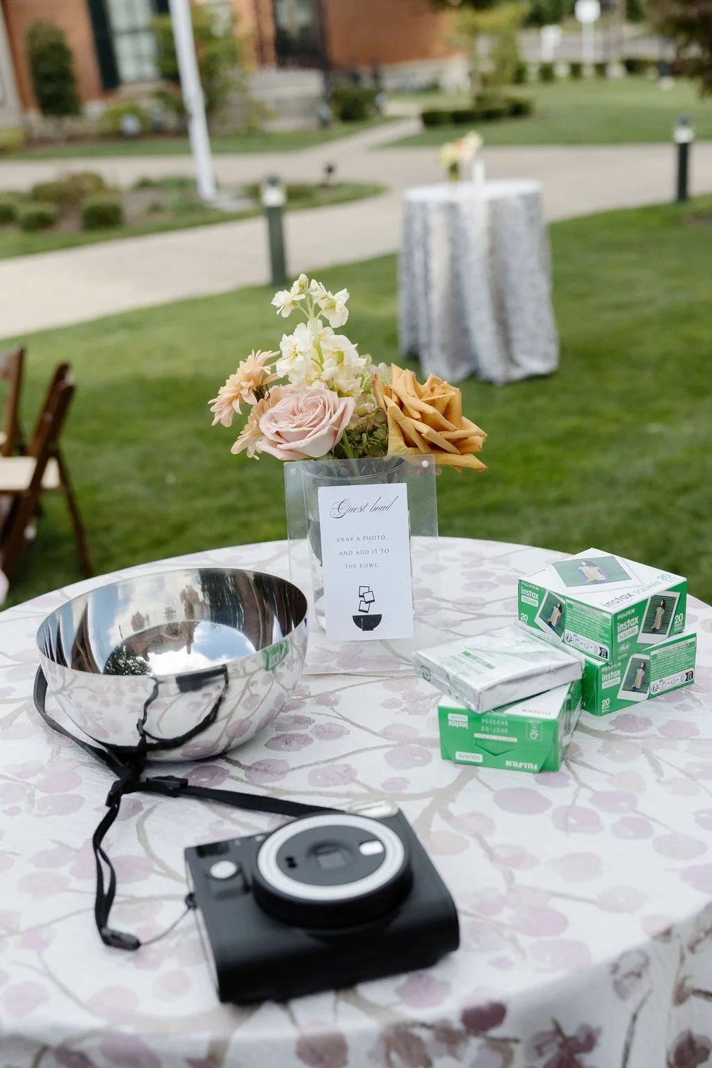 An outdoor event table decorated with a floral centerpiece, a camera, a metal bowl, and boxes of instant film.