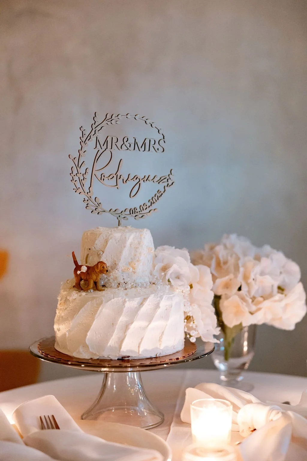 A white wedding cake with a 'Mr & Mrs Regal' cake topper, a small dog figurine, and white flower decorations, displayed on a glass cake stand with a floral centerpiece and candles around it.