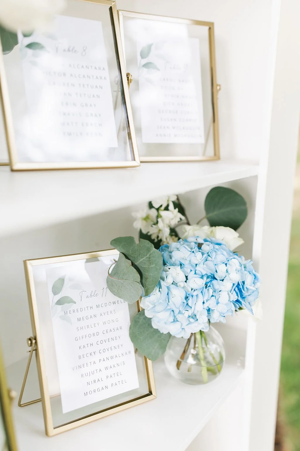 Names of guests on table cards on a white shelf, with a blue hydrangea flower in a glass vase and greenery.