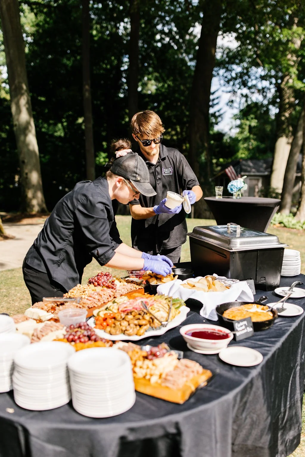 Two catering workers preparing food at an outdoor event with a buffet table laden with various dishes, grapes, and desserts, set against a backdrop of trees and outdoor tables.