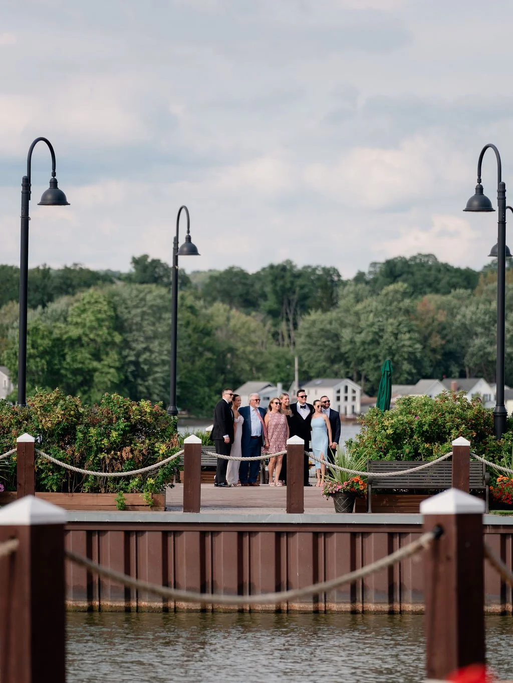 Group of people dressed in formal attire standing on a wooden dock near a body of water, with lush trees and houses in the background, under a partly cloudy sky.