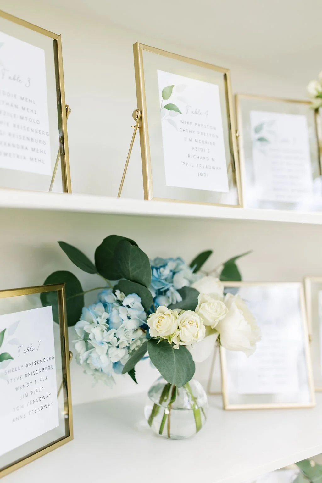 Table plan signs with gold frames and a floral centerpiece with white roses, blue hydrangeas, and greenery.