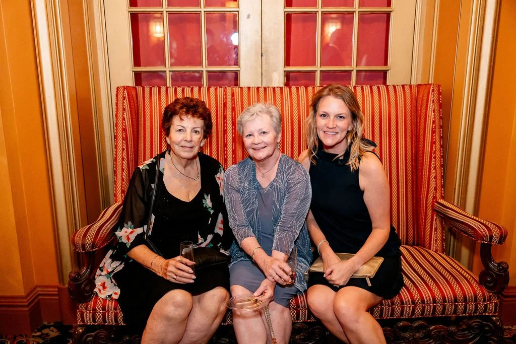 Three women sitting on a red and gold patterned vintage sofa in a warmly lit room with gold walls and a glass-paned wooden double door behind them.
