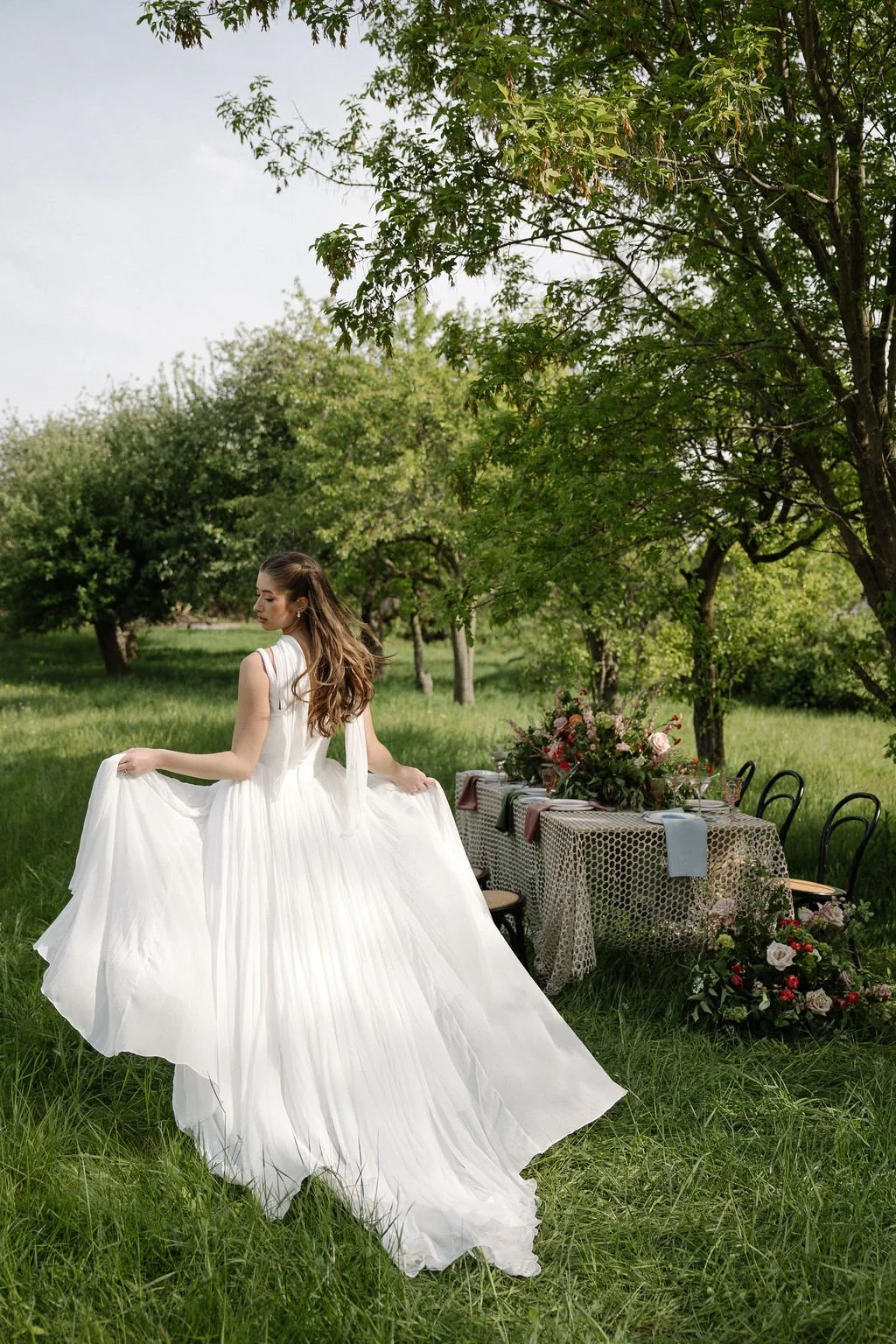 A woman in a white flowing dress standing in a lush green outdoor setting with a decorated table and floral arrangements.
