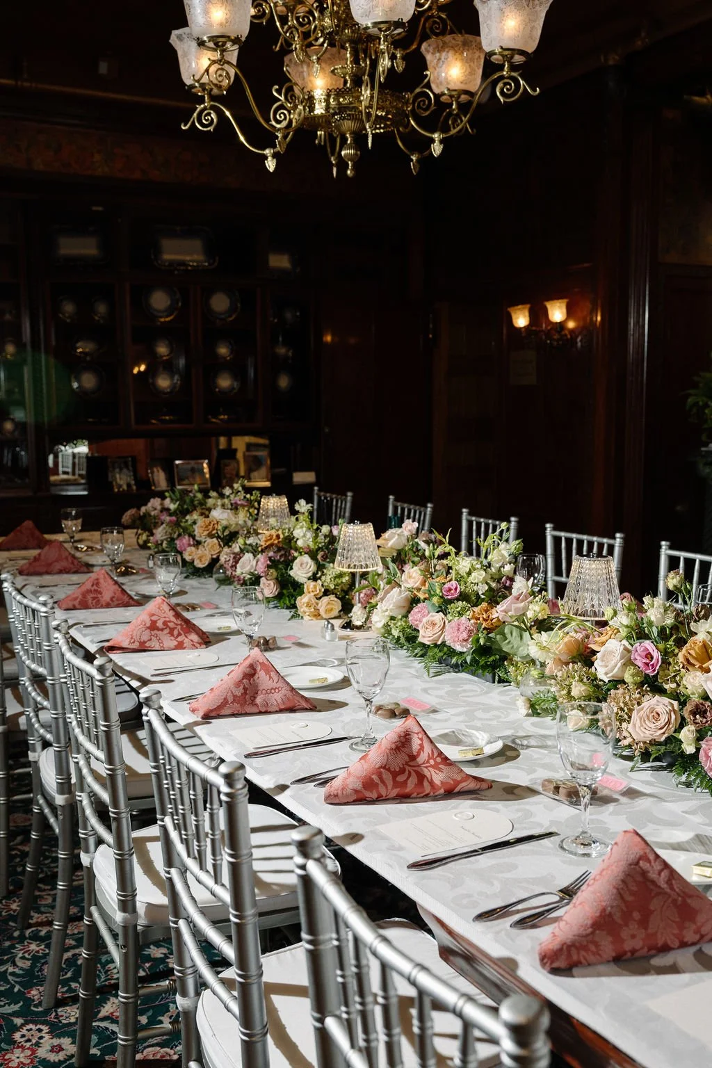 Elegant dining table set for a formal event, decorated with a lush floral centerpiece, pink napkins, and glassware under a chandelier.