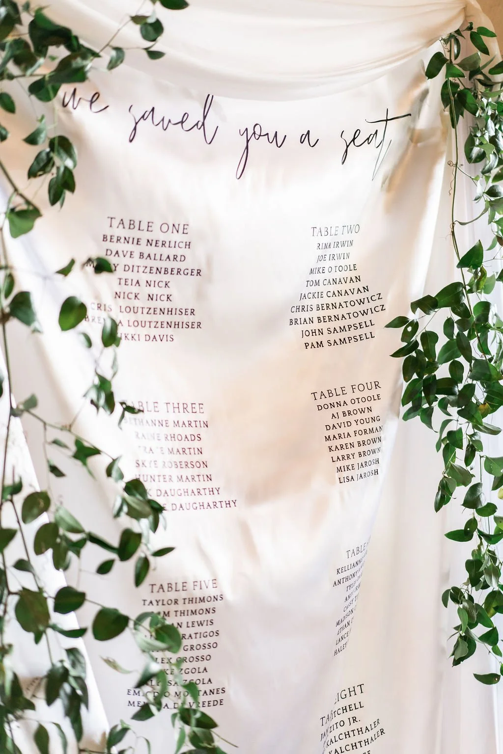 Wedding seating chart with the phrase "we saved you a seat" at the top, surrounded by green leafy foliage. The chart lists names grouped by tables, including tables one through five.