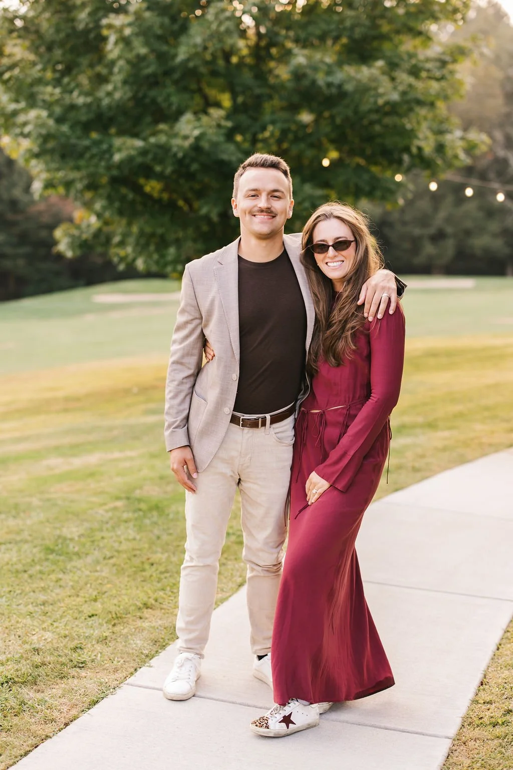 A smiling man and woman standing on a sidewalk outdoors, with green trees in the background, embracing each other. The man wears a light gray blazer, black shirt, and beige pants. The woman wears a long burgundy dress, sunglasses, and white sneakers 