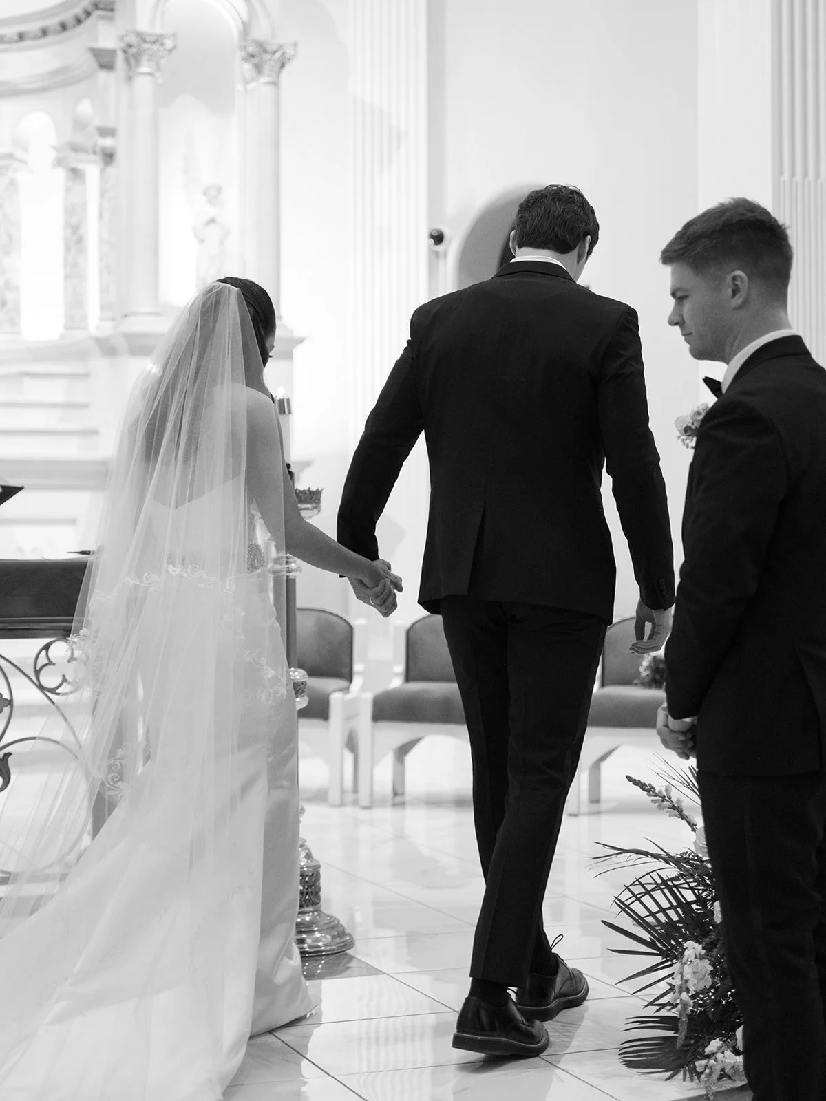 Black and white photo of a wedding ceremony, with bride and groom holding hands, while a groomsman stands nearby, in an indoor church or chapel setting.