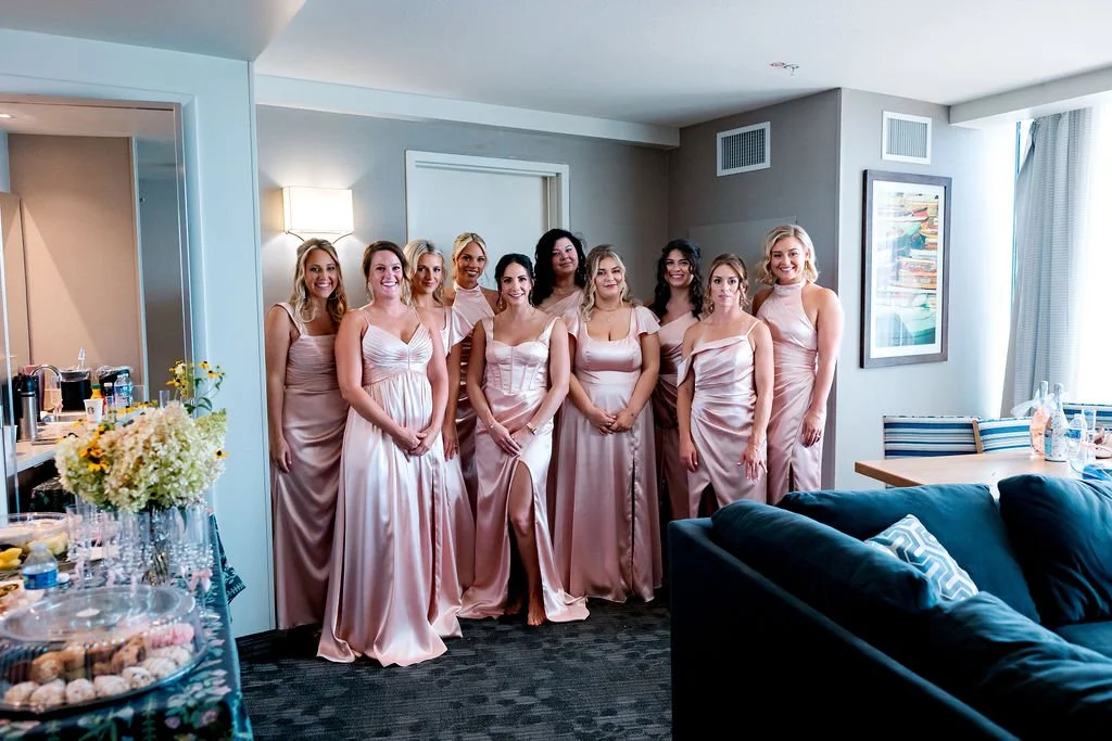 Ten women in matching satin dresses posing together in a hotel room with bright natural light