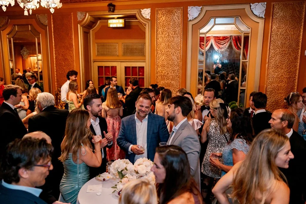 Guests socializing at a formal event in an ornate decorated ballroom with large mirrors and chandeliers.