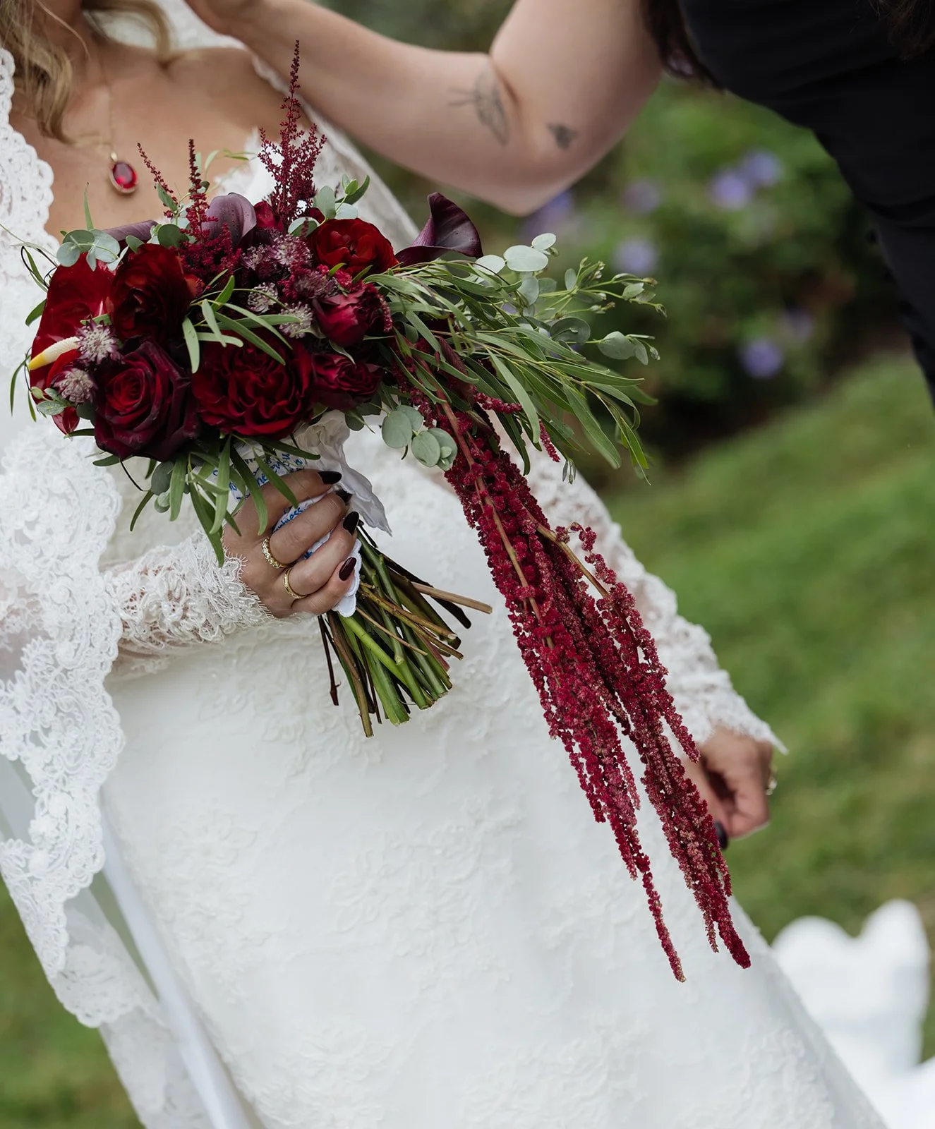 Close-up of a bride holding a vibrant bouquet of deep red and burgundy flowers, accented with greenery, in an outdoor setting with blurred greenery in the background.