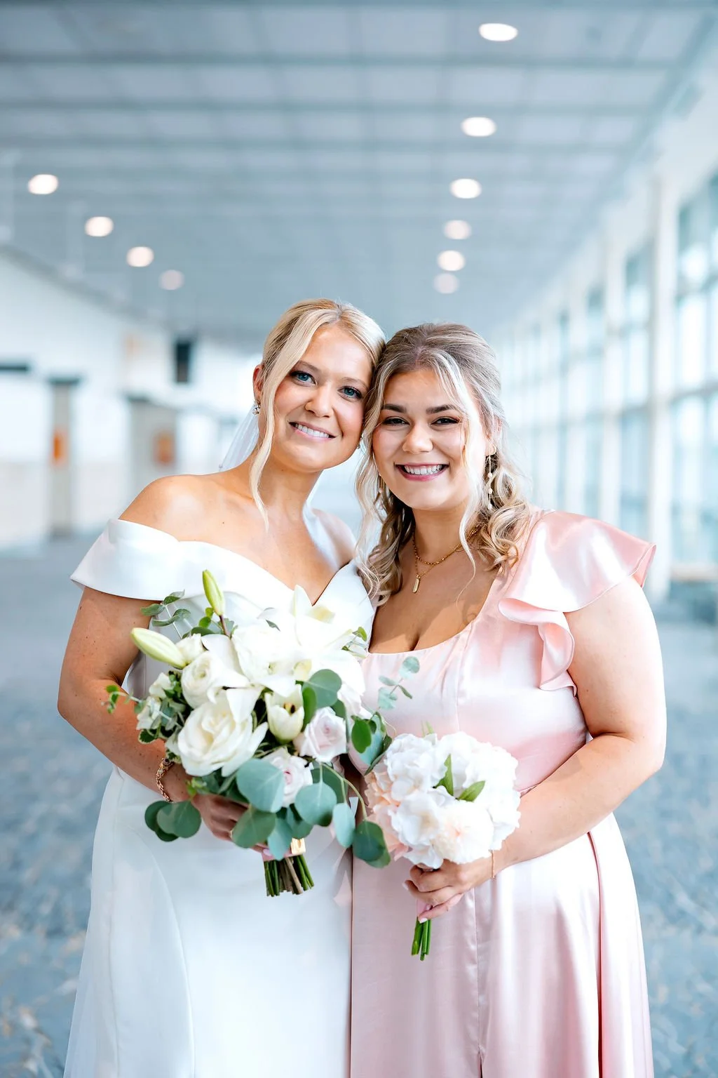 Two smiling women, one in a white wedding gown holding a bouquet of white flowers, and the other in a pink satin dress holding a bouquet, standing in a bright, modern building with large windows.