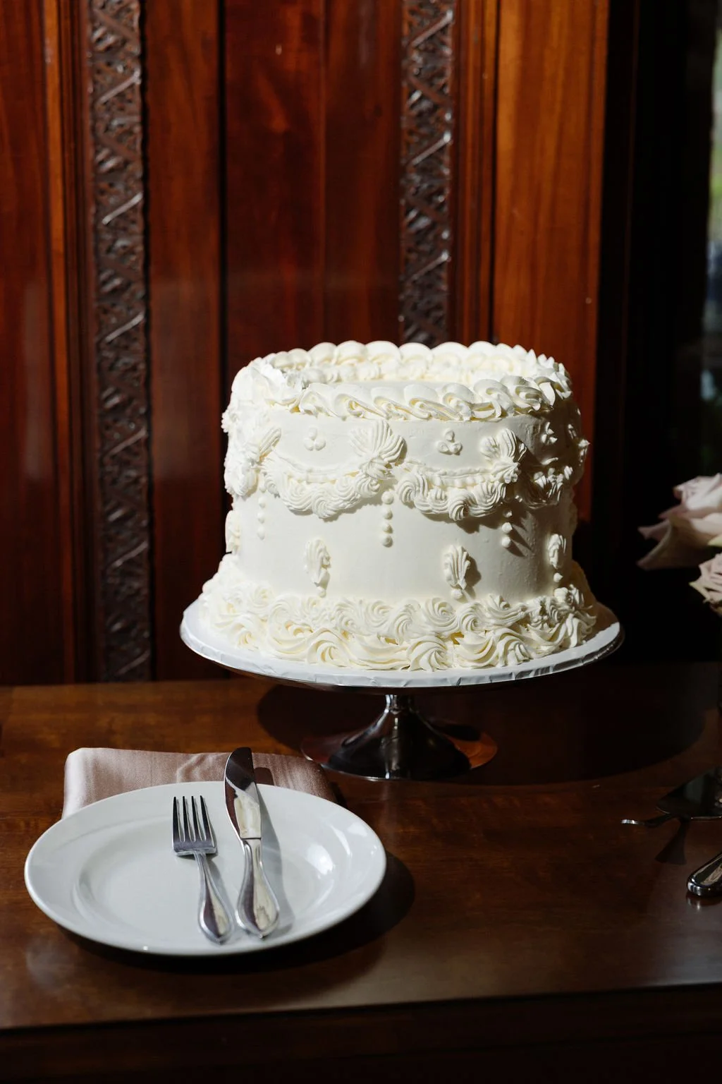 A white, elaborately decorated wedding or celebration cake with piped frosting and shell decorations, placed on a stand on a wooden table, with a plate, fork, and knife in front of it.