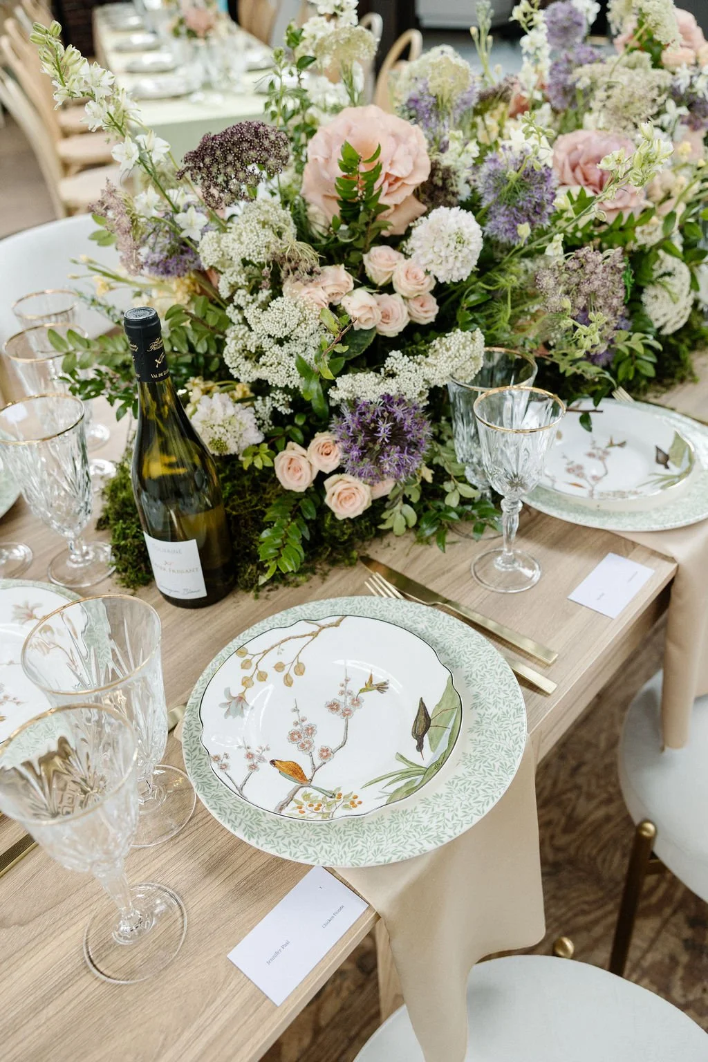 Elegant table setting with a large floral centerpiece featuring pink, white, and purple flowers. There are patterned plates, crystal glasses, gold flatware, a wine bottle, and place cards on a light-colored wooden table.