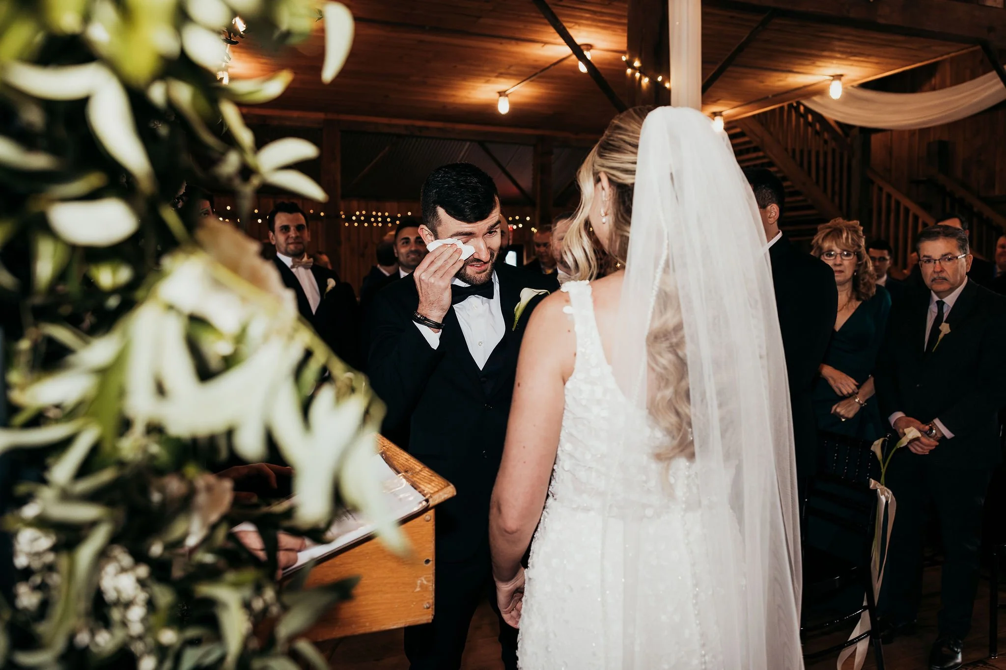 A groom wiping tears during his wedding ceremony as the bride stands in front of him. Guests look on inside a wooden venue with string lights.