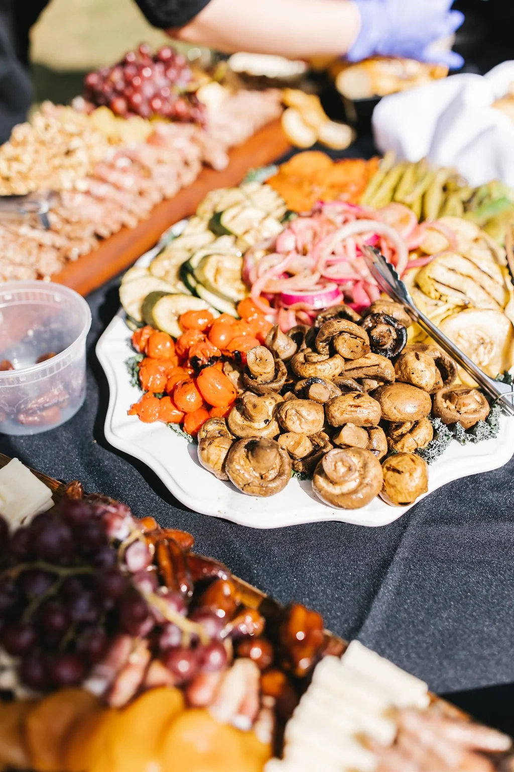 Assorted grilled vegetables on a white platter, including mushrooms, cherry tomatoes, zucchini, pickled onions, and roasted carrots, at a food buffet.