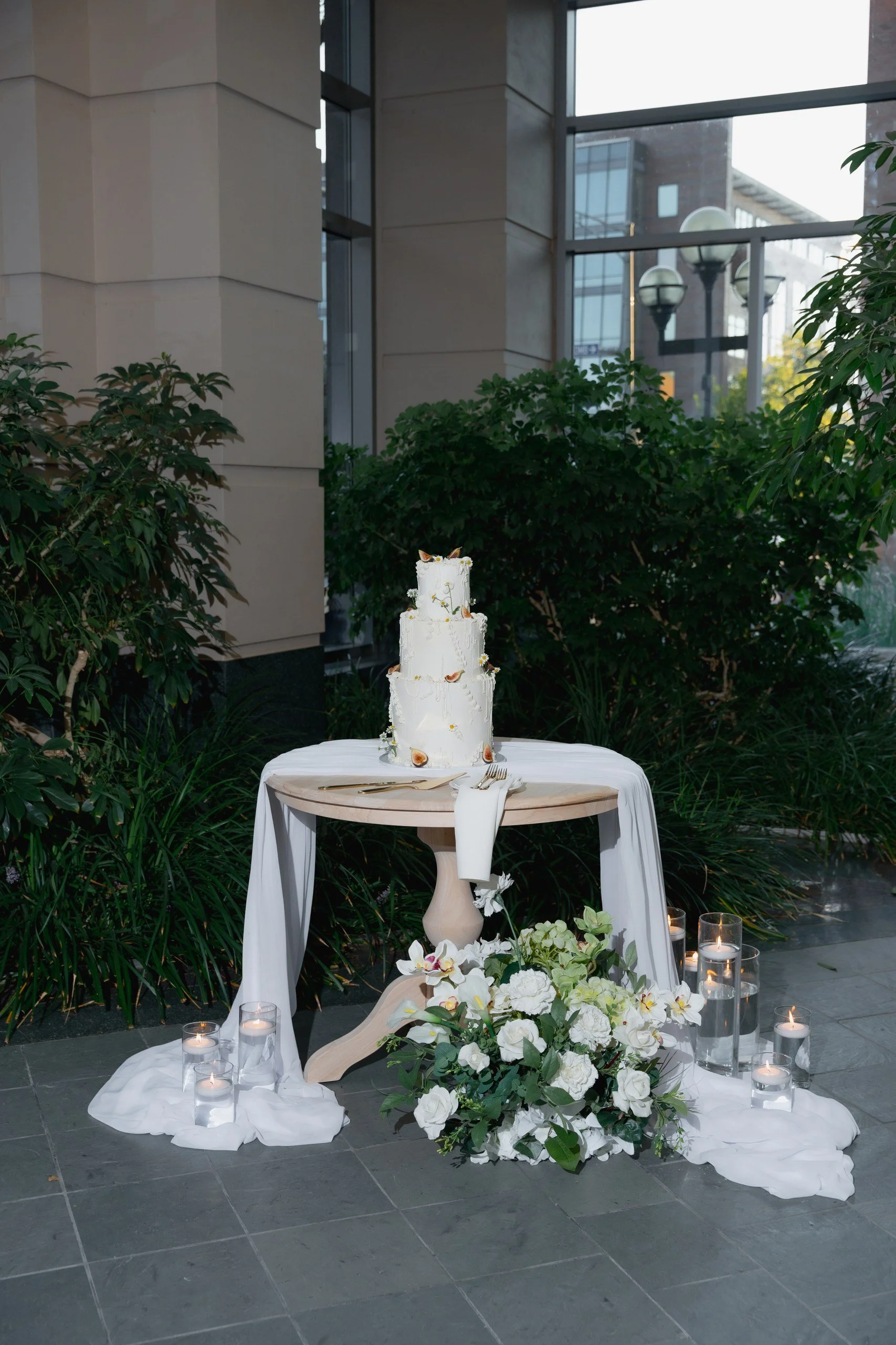 A white wedding cake with floral decorations on a round table draped with white fabric, surrounded by white flowers and lit candles in glass holders, set against green plants inside a building with large windows.