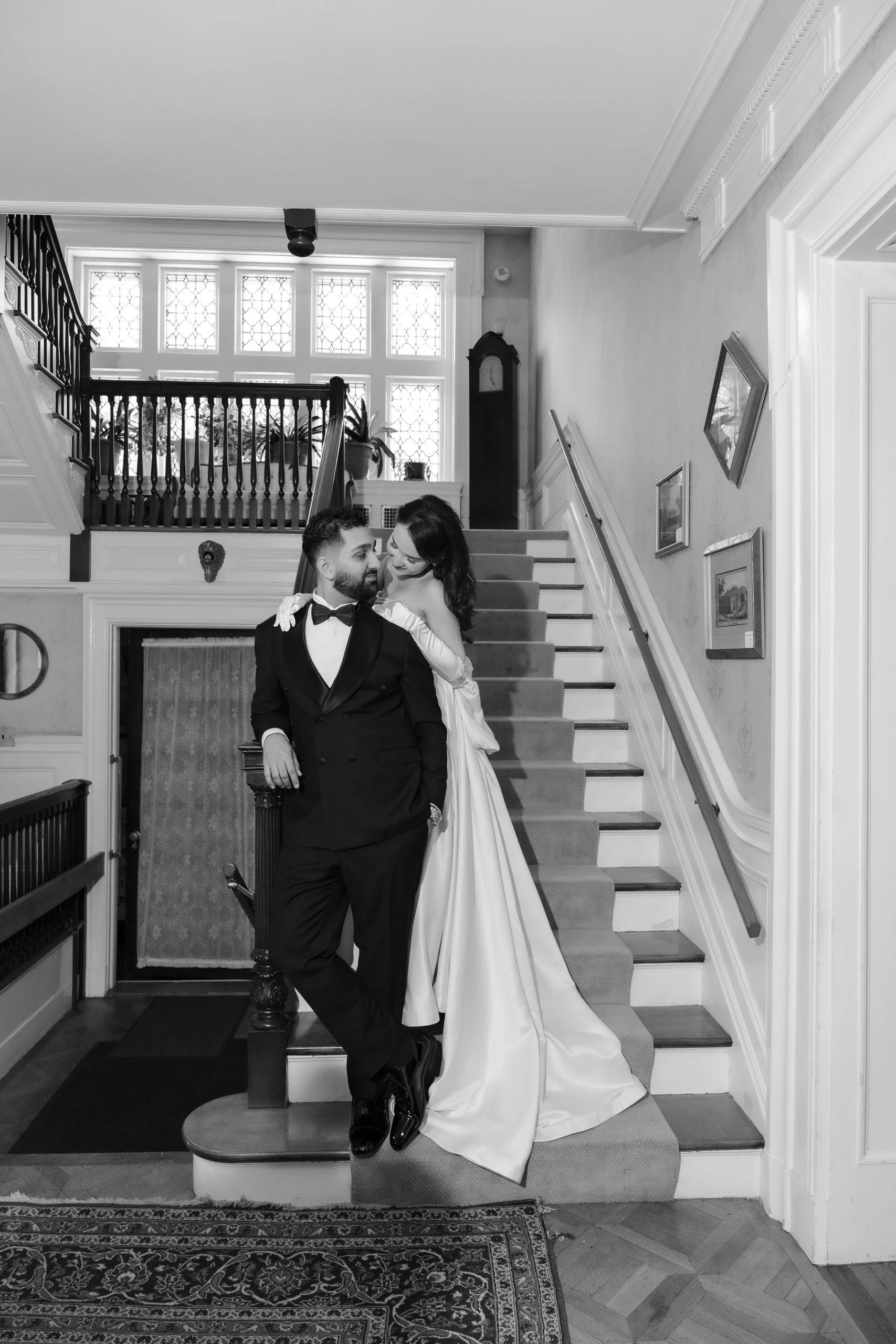 A black and white photo of a couple in formal attire standing on a staircase inside a house. The man is wearing a tuxedo and the woman is in an elegant white gown. They are close together, smiling and looking at each other.
