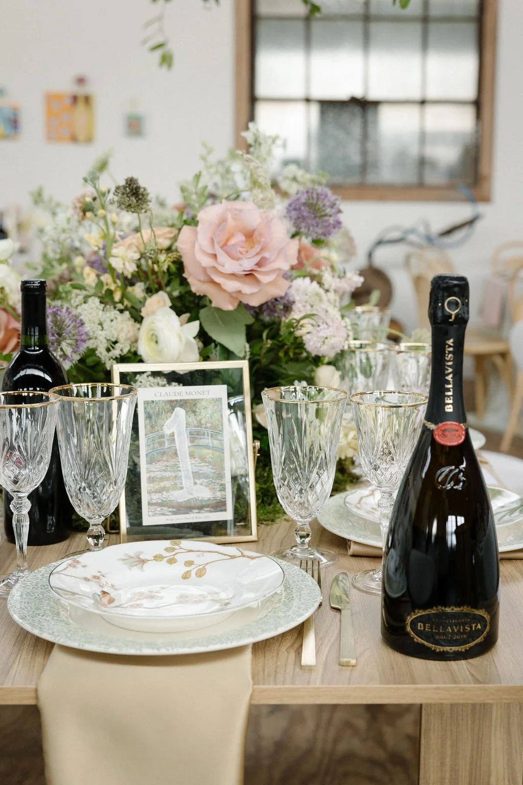 A beautifully set dining table with floral centerpiece, wine bottles, glassware, plates, and cutlery, in a bright, rustic room with a window.