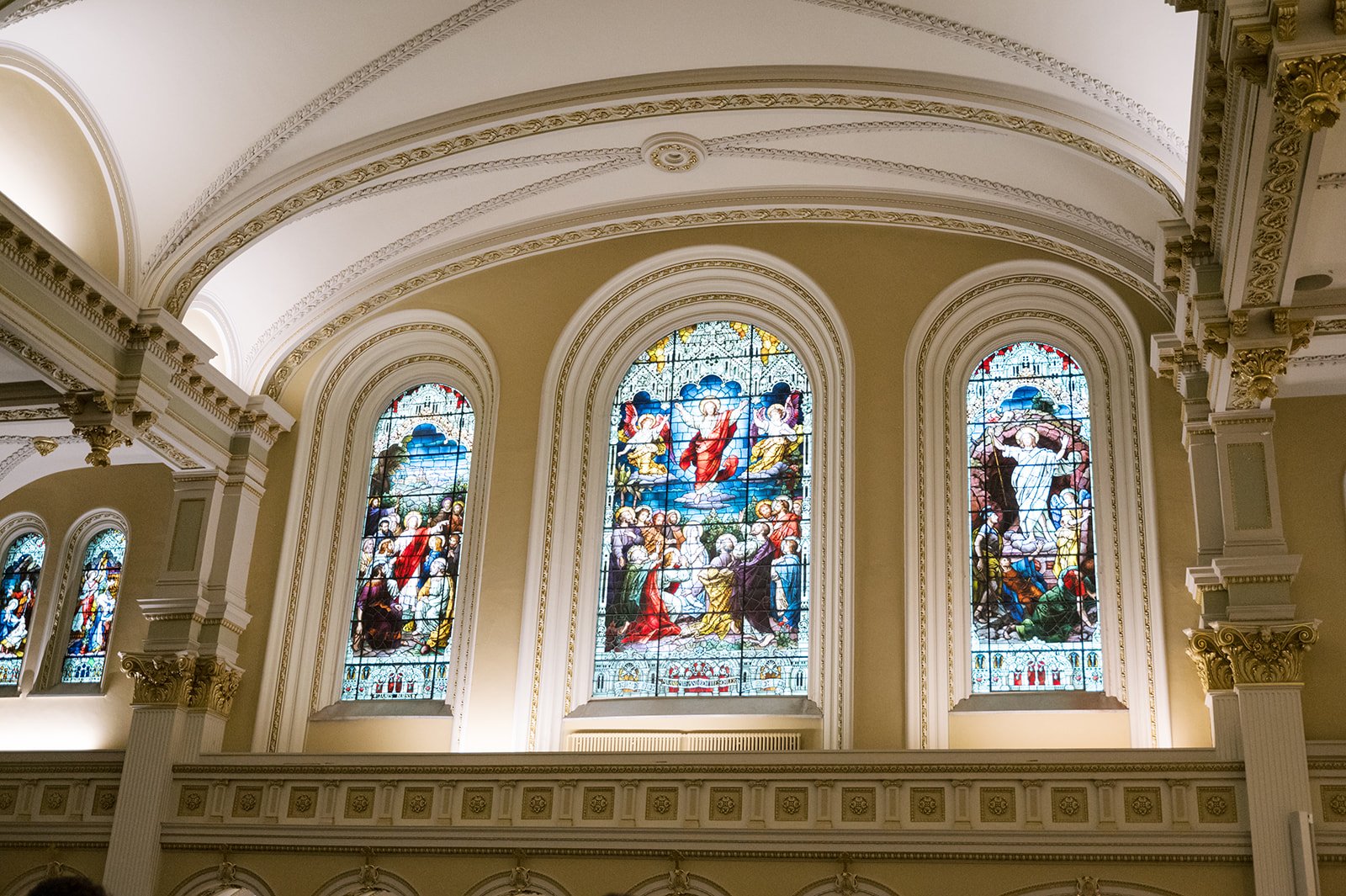 Inside a church with ornate white and gold architectural details and stained glass windows depicting religious scenes.