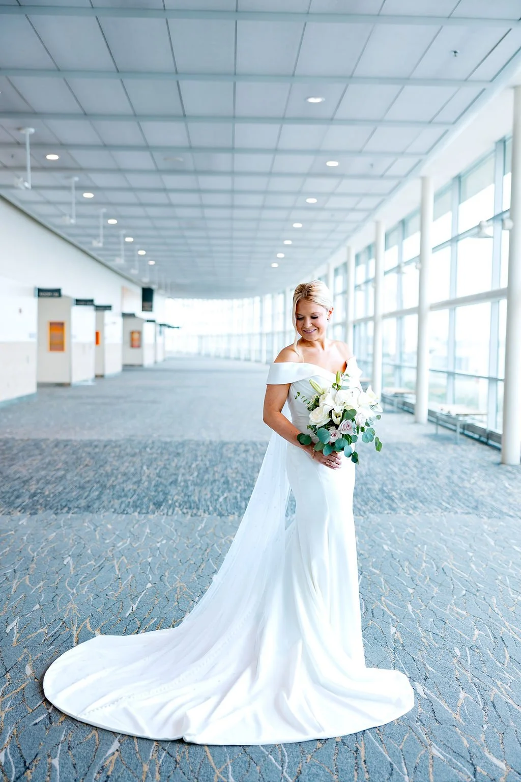 Bride in a white off-the-shoulder wedding gown holding a bouquet of white and pale pink flowers inside an airport terminal.