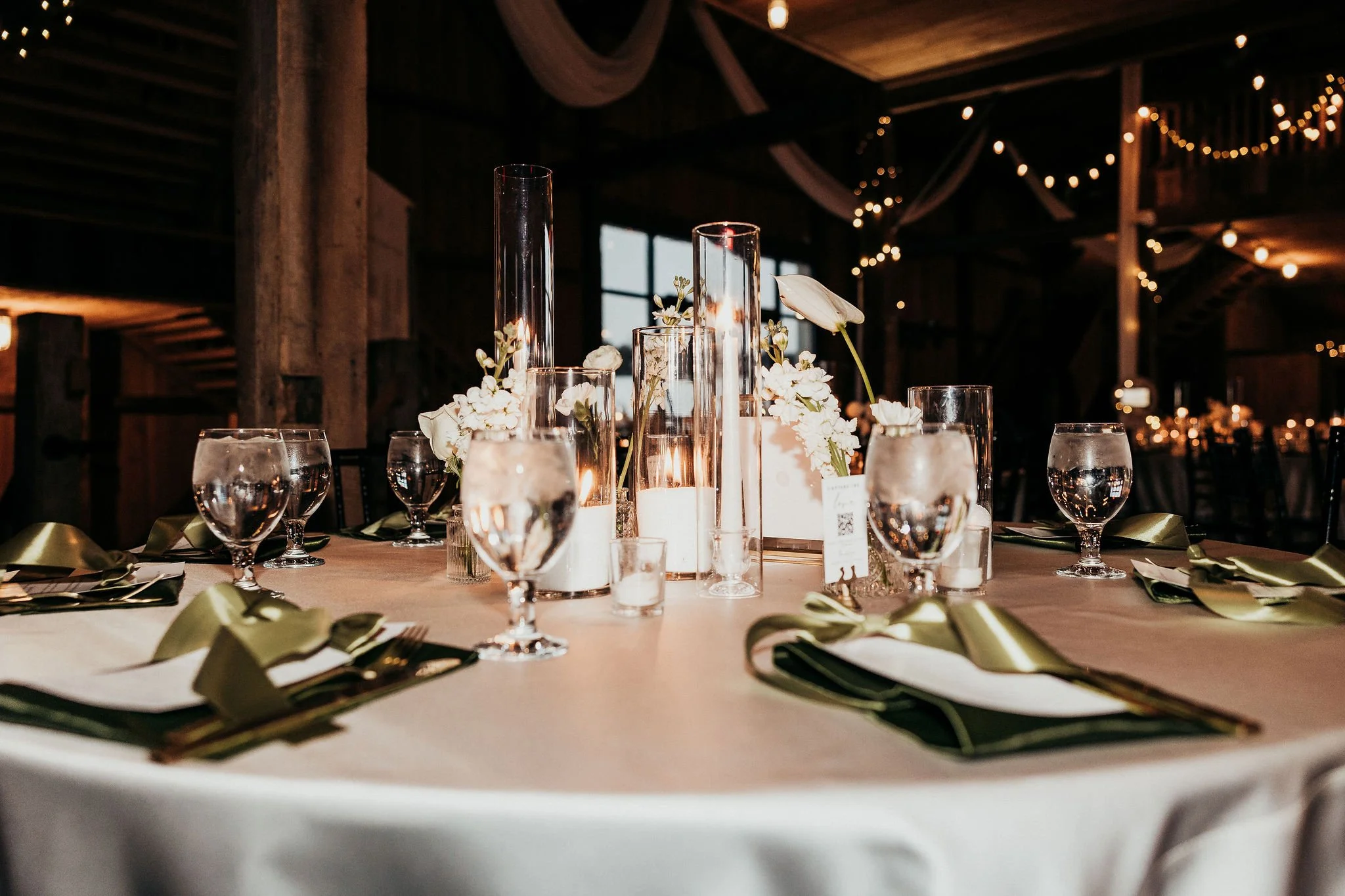 A decorated banquet table set with glasses, candles, and floral centerpieces at an elegant event venue with string lights and wooden beams.